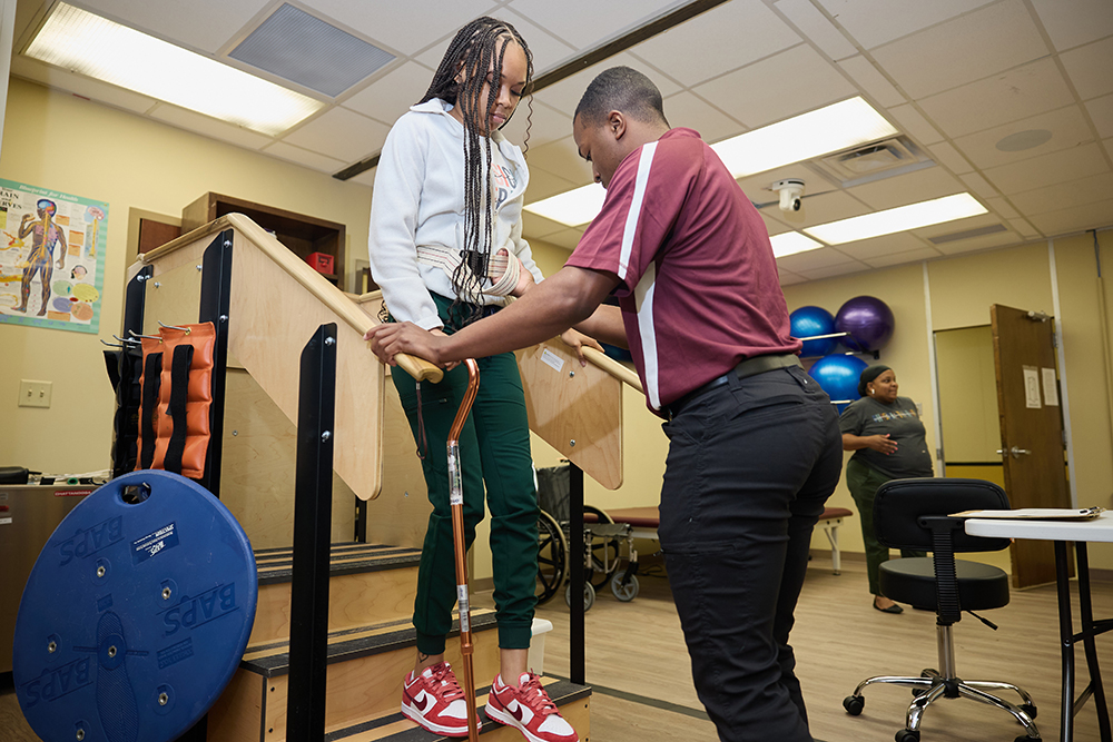 Students practice in a lab. A woman holding a cane navigates a staircase while a student guides her down.