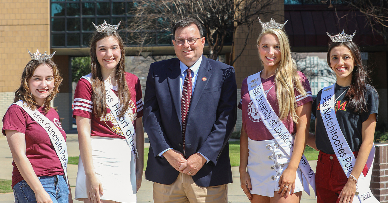 Five people smile at the camera. In the middle is a man with a suit. On either side are two women wearing sashes and crowns.