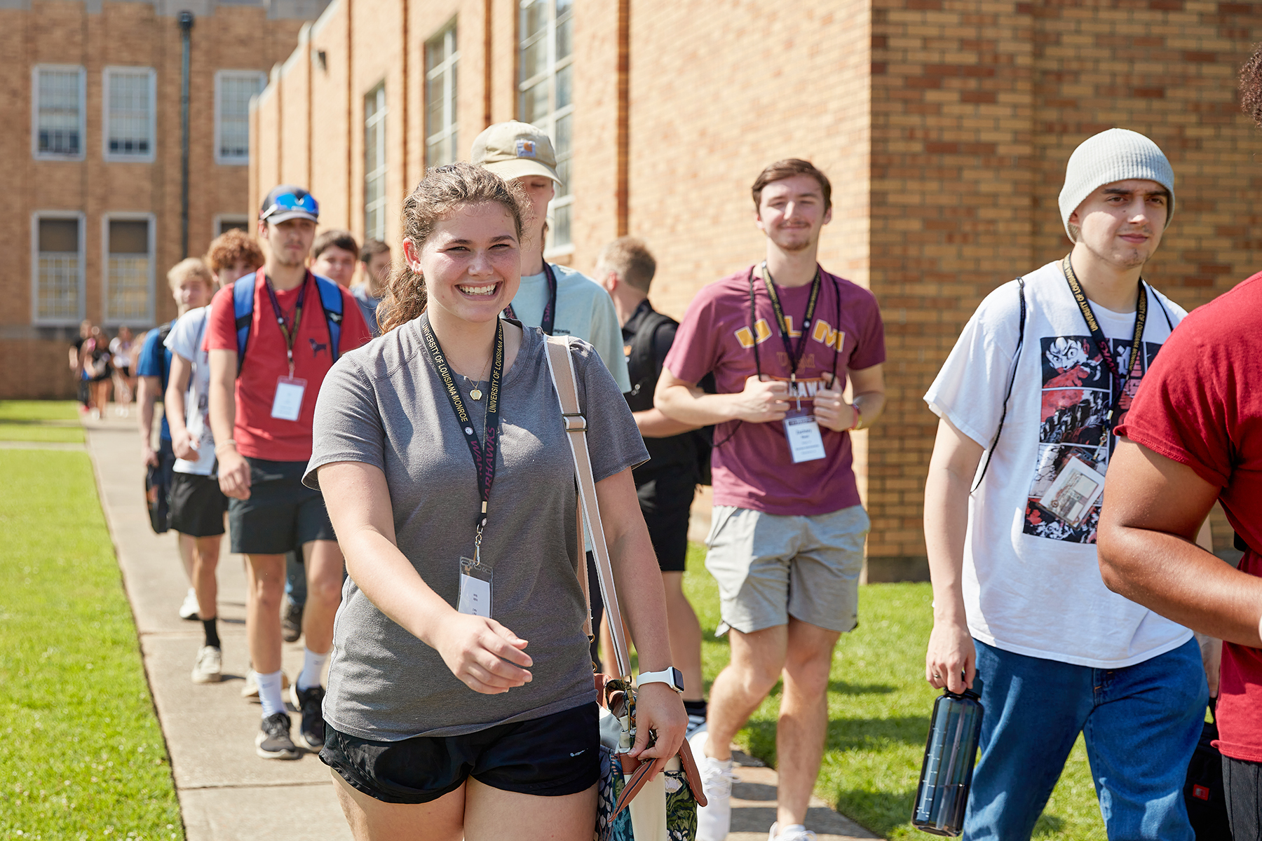 A group of young people walk on a side walk in front of a brick building. A woman in front smiles at the camera.